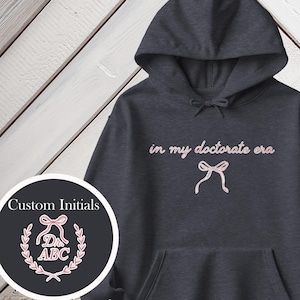 Könnte beinhalten: Dunkelgrauer Kapuzenpullover mit dem Schriftzug "in my doctorate era" in rosa Schrift. Darunter befindet sich eine rosa Schleife. Der Hoodie hat eine Vordertasche und eine Kapuze. Auf der linken Seite befindet sich eine runde Grafik mit individuellen Initialen.