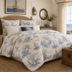 May include: A bedroom with a nautical theme, featuring a bed with a blue and white toile design of sailboats and shells. The room includes a woven headboard, bedside table, and a woven trunk. A painting of a sailing boat hangs above the bed, with a sea view visible.