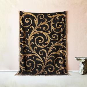 May include: Black throw blanket with a gold, swirling, baroque-style pattern. The blanket has a fringed edge and is displayed against a pink and white background. The design features ornate, floral-like elements.