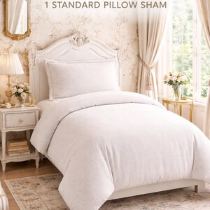 Puede incluir: Un juego de cama Twin XL con un edred&oacute;n y una funda de almohada est&aacute;ndar. El juego es de color gris claro y se muestra en una estructura de cama blanca. El fondo incluye un papel pintado con motivos florales, un espejo dorado y una mesita de noche.
