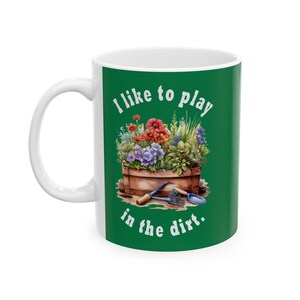 Könnte beinhalten: Weiße Keramiktasse mit grünem Hintergrund und dem Text "Ich spiele gerne im Dreck". Ein Blumentopf mit bunten Blumen und Gartengeräten ist abgebildet.
