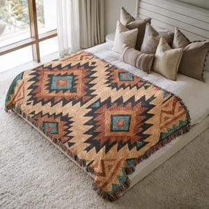 May include: A woven blanket with a geometric pattern in shades of tan, brown, black, and teal. The blanket is rectangular and has a fringed edge. The design features repeating diamond and zigzag shapes, creating a Southwestern or tribal aesthetic.