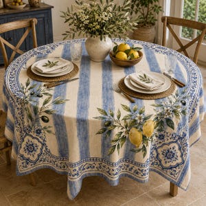 Puede incluir: Un mantel redondo con rayas verticales azules y crema, decorado con ramas de olivo, limones y patrones de azulejos florales azules. La tela está sobre una mesa redonda con dos sillas de madera, platos blancos y un cuenco de limones.