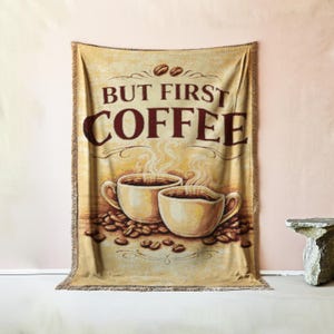 Op de afbeelding: Een beige sprei met de tekst "BUT FIRST COFFEE" in bruin. De sprei heeft twee koffiekopjes, koffiebonen en stoom die uit de kopjes opstijgt. De sprei heeft een gefranjerde rand.