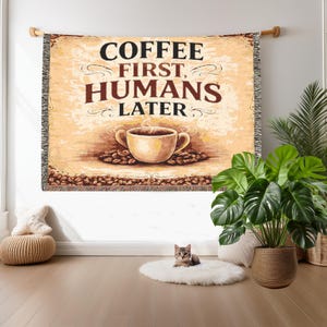 Könnte beinhalten: Gewebte Wandtapete mit dem Text "COFFEE FIRST, HUMANS LATER" in Schwarz und Braun. Die Tapete zeigt eine Kaffeetasse und Kaffeebohnen. Die Tapete hängt an einer Holzstange.