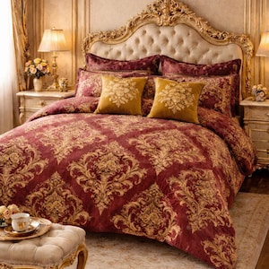 May include: A luxurious bedroom setting featuring a bed with a tufted headboard and ornate gold trim. The bedding includes a burgundy comforter and pillows with a gold damask pattern. Two gold throw pillows with floral designs add a touch of elegance.