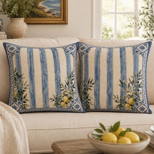 May include: Two square throw pillows featuring a cream-colored base with vertical blue stripes. The design includes a decorative blue and white tile-patterned border and a printed illustration of green olive branches and yellow lemons.