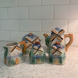 May include: A vintage tea set with a windmill design. The set includes a teapot, creamer, sugar bowl, and two cups. The pieces are made of ceramic and are painted in shades of blue, green, and brown.