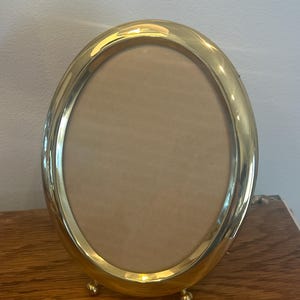 May include: An oval-shaped picture frame with a polished gold-tone metal finish. The frame features a smooth, rounded border and is supported by two small, spherical feet at the base, allowing it to stand upright on a flat surface.