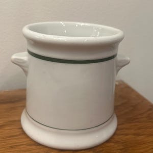 May include: White ceramic shaving mug with a green stripe around the middle and base. The mug has two small handles and a flared base. The mug is sitting on a wooden surface.