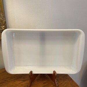 May include: A rectangular, white ceramic baking dish with rounded corners, displayed on a small, wooden stand. The dish is empty and clean, reflecting the light. The stand is a dark brown color.
