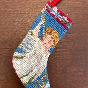May include: A small, blue and red Christmas stocking with an embroidered image of an angel holding a string of lights. The stocking has a red felt loop at the top.