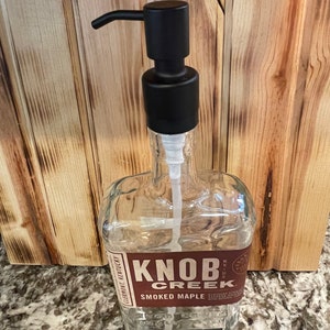 May include: A clear glass bottle repurposed as a soap dispenser, featuring a black pump and the "Knob Creek Smoked Maple" label. The bottle is set against a wooden background.