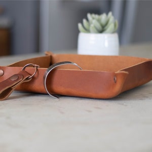 May include: A brown leather tray with a key chain and a metal bracelet inside. The tray has a square shape with rounded corners and is made of a soft, supple leather. The tray is perfect for holding small items like keys, jewelry, or change.