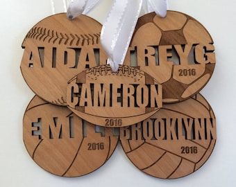 May include: Four wooden ornaments with different sports themes. Each ornament features a different sport: baseball, football, soccer, and volleyball. Each ornament has a name and the year 2016 engraved on it. The names are Aidan, Cameron, Trey, and Brooklynn.