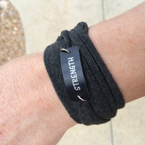 Puede incluir: Una pulsera de tela gris con una etiqueta de metal negro que dice "STRENGTH" en letras blancas.