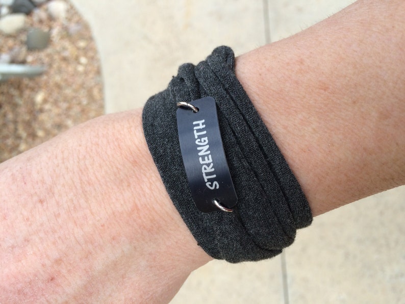 Puede incluir: Una pulsera de tela gris con una etiqueta de metal negro que dice "STRENGTH" en letras blancas.