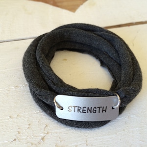 Puede incluir: Un brazalete de tela gris con una etiqueta plateada que dice "STRENGTH".
