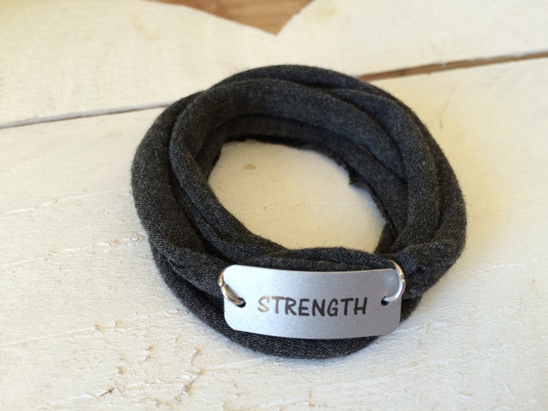 Puede incluir: Un brazalete de tela gris con una etiqueta plateada que dice "STRENGTH".