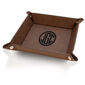 May include: A brown leather valet tray with a personalized monogram in a black circle. The tray has folded corners secured with metal snaps. The interior is a lighter shade of brown than the exterior.