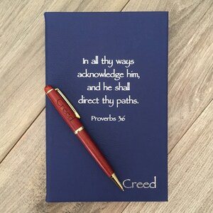 May include: A blue leather-bound journal with a gold trim and the inscription "In all thy ways acknowledge him, and he shall direct thy paths. Proverbs 3:6"  The journal is accompanied by a wooden pen with the name "Creed" engraved on it.