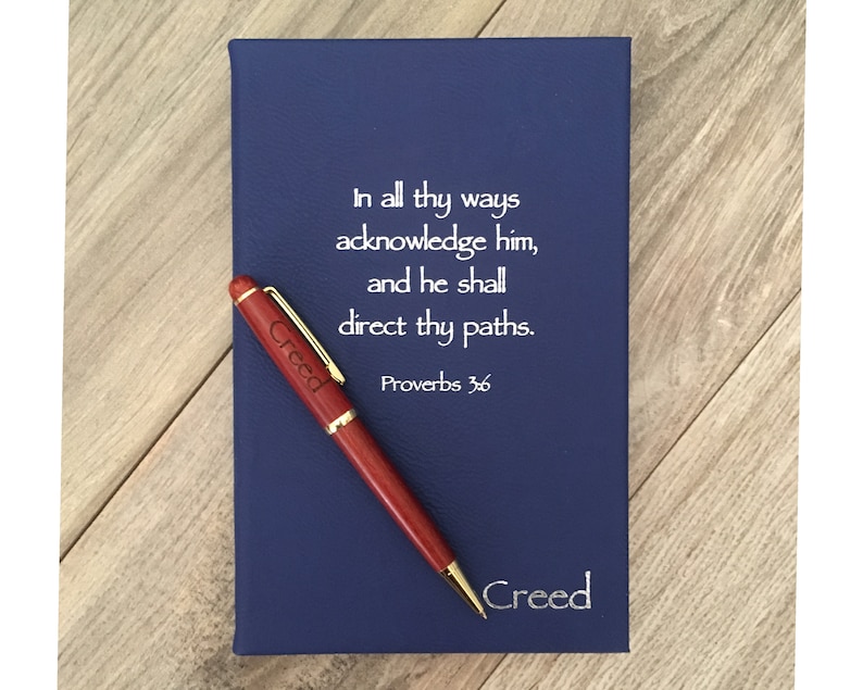 May include: A blue leather-bound journal with a gold trim and the inscription "In all thy ways acknowledge him, and he shall direct thy paths. Proverbs 3:6"  The journal is accompanied by a wooden pen with the name "Creed" engraved on it.