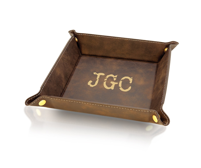 May include: A brown leather valet tray with the initials "JGC" in gold lettering. The tray has a stitched border and gold-colored snap closures at each corner. The interior is a lighter shade of brown.