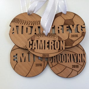 May include: Four wooden ornaments shaped like different sports balls, each with a name and the year 2016 engraved on them. The ornaments are baseball, soccer, football, and volleyball.
