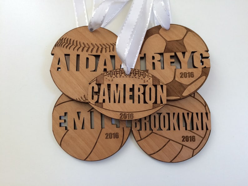 May include: Four wooden ornaments shaped like different sports balls, each with a name and the year 2016 engraved on them. The ornaments are baseball, soccer, football, and volleyball.