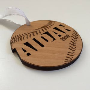 May include: A wooden baseball ornament with the name "Aidan" and the year "2016" engraved on it. The ornament has a white ribbon attached for hanging.