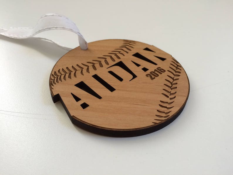 May include: A wooden baseball ornament with the name "Aidan" and the year "2016" engraved on it. The ornament has a white ribbon attached for hanging.