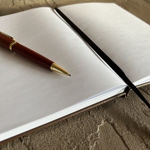 May include: A brown leather bound journal with a black ribbon bookmark. The journal is open to a blank page with a gold pen lying on the page.