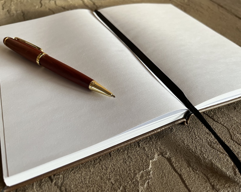May include: A brown leather bound journal with a black ribbon bookmark. The journal is open to a blank page with a gold pen lying on the page.