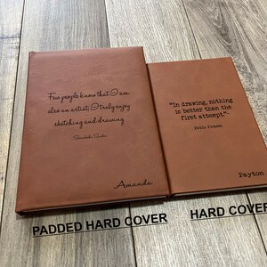 May include: Two brown leather-bound journals with quotes about art and drawing. The journal on the left has the quote "Few people know that I am also an artist, truly enjoy sketching and drawing" by Susiske Sake and the name "Amanda" inscribed on the cover. The journal on the right has the quote "In drawing, nothing is better than the first attempt" by Pablo Picasso and the name "Payton" inscribed on the cover. The journal on the left is labeled "PADDED HARD COVER" and the journal on the right is labeled "HARD COVER".