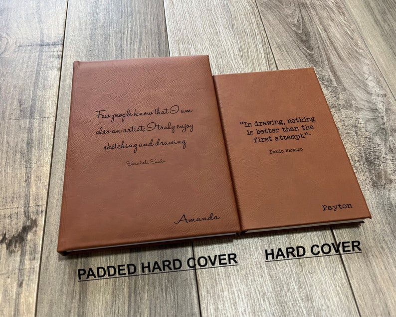 May include: Two brown leather-bound journals with quotes about art and drawing. The journal on the left has the quote "Few people know that I am also an artist, truly enjoy sketching and drawing" by Susiske Sake and the name "Amanda" inscribed on the cover. The journal on the right has the quote "In drawing, nothing is better than the first attempt" by Pablo Picasso and the name "Payton" inscribed on the cover. The journal on the left is labeled "PADDED HARD COVER" and the journal on the right is labeled "HARD COVER".