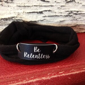 Puede incluir: Pulsera de tela negra con una etiqueta de metal plateado que dice "Be Relentless".