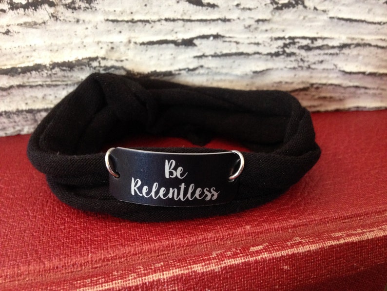 Puede incluir: Pulsera de tela negra con una etiqueta de metal plateado que dice "Be Relentless".