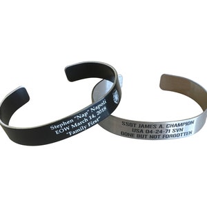 May include: Two engraved metal bracelets. The black bracelet reads "Stephen "Nap" Napoli EOW March 14, 2018 "Family First". The silver bracelet reads "SSGT JAMES A. CHAMPION USA 04-24-71 SVN GONE BUT NOT FORGOTTEN".