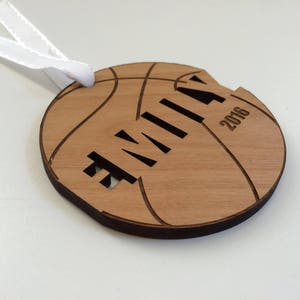 May include: A wooden basketball ornament with the name "Emily" and the year "2016" engraved on it. The ornament has a white ribbon attached to it.