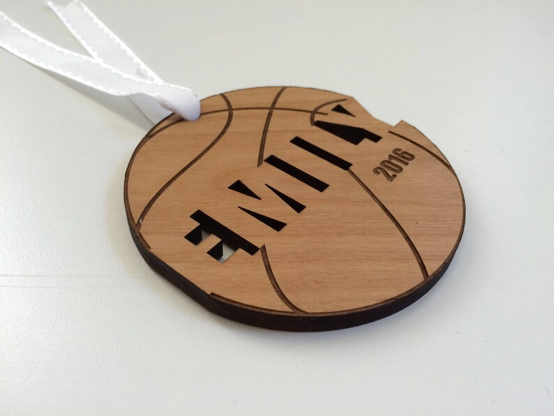 May include: A wooden basketball ornament with the name "Emily" and the year "2016" engraved on it. The ornament has a white ribbon attached to it.