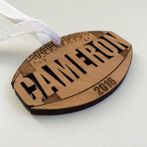 May include: A wooden football ornament with the name "CAMERON" and the year "2016" engraved on it. The ornament has a white ribbon attached to it.
