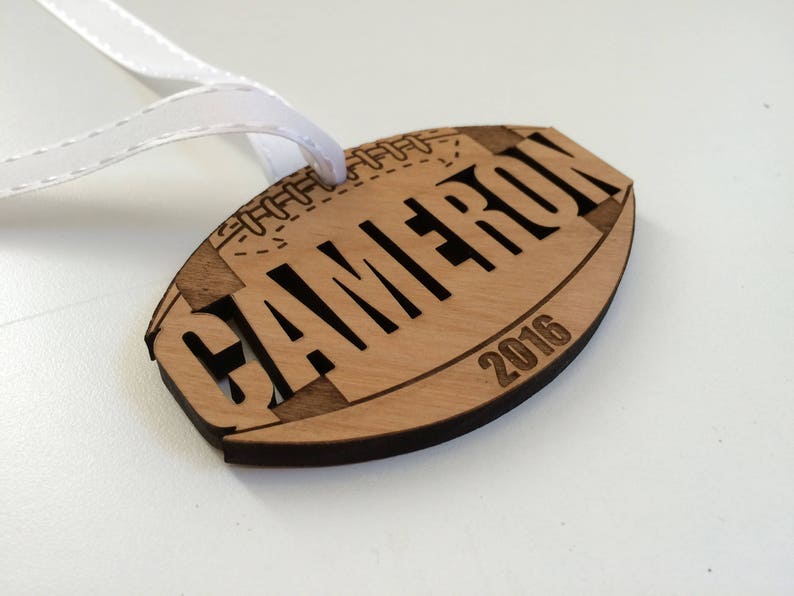 May include: A wooden football ornament with the name "CAMERON" and the year "2016" engraved on it. The ornament has a white ribbon attached to it.