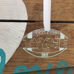 Pu&ograve; includere: Un ornamento da albero di Natale a forma di pallone da calcio in acrilico bianco con il nome "Tyce" e l'anno "2017" incisi su di esso. L'ornamento &egrave; appeso a un nastro bianco.
