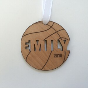 May include: A wooden basketball ornament with the name "EMILY" cut out and the year "2016" inscribed below. The ornament is brown and has a white ribbon attached.