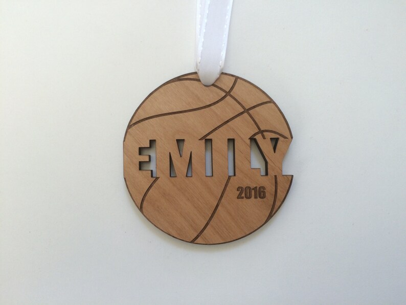 May include: A wooden basketball ornament with the name "EMILY" cut out and the year "2016" inscribed below. The ornament is brown and has a white ribbon attached.