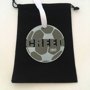 Pu&ograve; includere: Un ornamento a forma di pallone da calcio bianco e grigio con la parola "GREEF" e l'anno "2017" stampati su di esso. L'ornamento &egrave; appeso a un nastro bianco e si trova in una borsa di velluto nero.