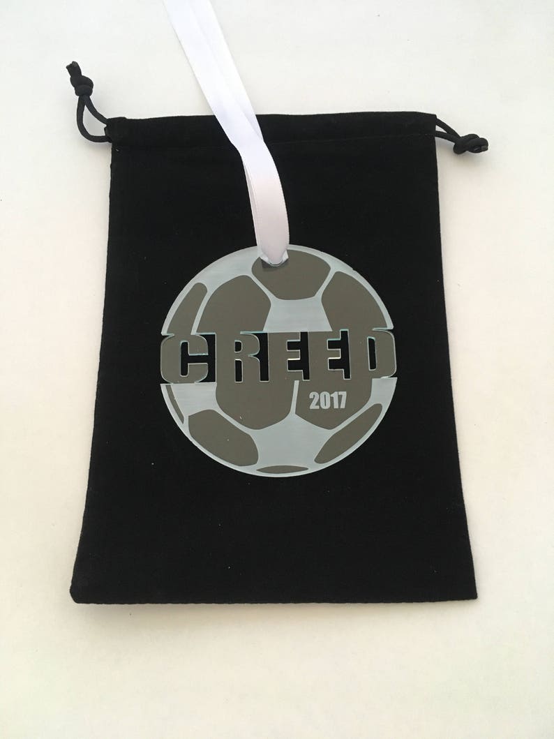 Pu&ograve; includere: Un ornamento a forma di pallone da calcio bianco e grigio con la parola "GREEF" e l'anno "2017" stampati su di esso. L'ornamento &egrave; appeso a un nastro bianco e si trova in una borsa di velluto nero.