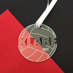 Pu&ograve; includere: Un ornamento da pallavolo bianco con il nome "KLARE" e l'anno "2017" inciso su di esso. L'ornamento &egrave; appeso a un nastro bianco su uno sfondo nero e rosso.