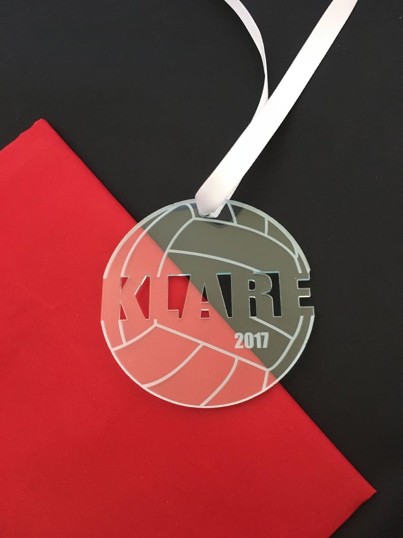 Pu&ograve; includere: Un ornamento da pallavolo bianco con il nome "KLARE" e l'anno "2017" inciso su di esso. L'ornamento &egrave; appeso a un nastro bianco su uno sfondo nero e rosso.
