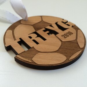 May include: A wooden soccer ball ornament with the name "TRAYCE" and the year "2016" engraved on it. The ornament is brown and has a white ribbon attached to it.